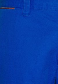 Detailní záběr na královsky modré kalhoty s poutkem na pásek a malým vyšívaným lemováním v červené, bílé a hnědé barvě poblíž kapsy.