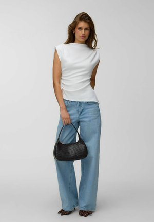 Kvinde iført hvid ærmeløs top og blå vidde ben jeans, der holder en sort flet håndtaske mod en ensfarvet baggrund.
