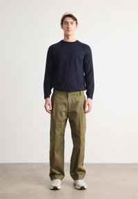 Mornarska majica z dolgimi rokavi, sproščen kroj, z logotipom; olivno zeleni hlače, ohlapen kroj; sivi športni copati z belimi detajli; model nosi svetlo kapo.