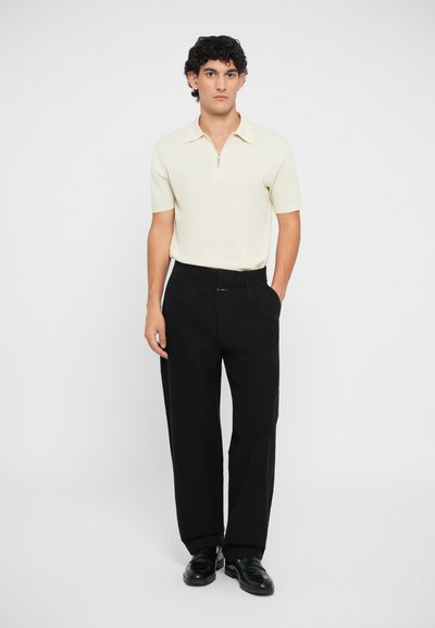 Polo de manga corta color beige claro con un cuello con cremallera, combinado con pantalones sueltos negros y zapatos negros. Diseño simple y limpio.