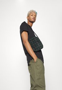 T-shirt preto, calças verde-oliva e uma bolsa cruzada em padrão xadrez verde. A bolsa possui um fecho de zip e um design texturizado.