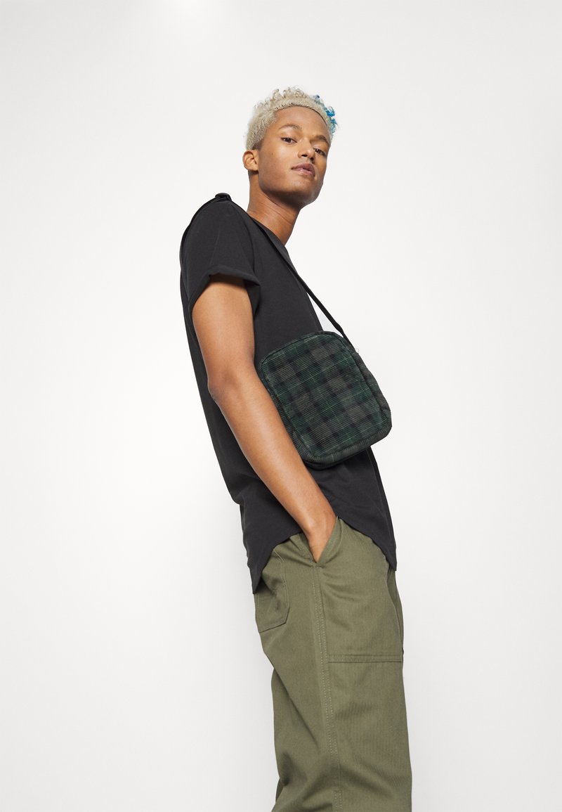 T-shirt preto, calças verde-oliva e uma bolsa cruzada em padrão xadrez verde. A bolsa possui um fecho de zip e um design texturizado.