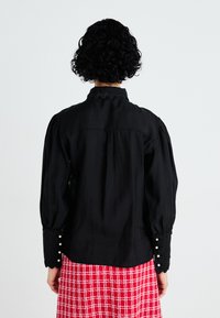 Blusa negra de manga larga con mangas abullonadas, cuello de scallop y botones en los puños; llevada sobre una falda de cuadros rojos. Textura de tela suave.