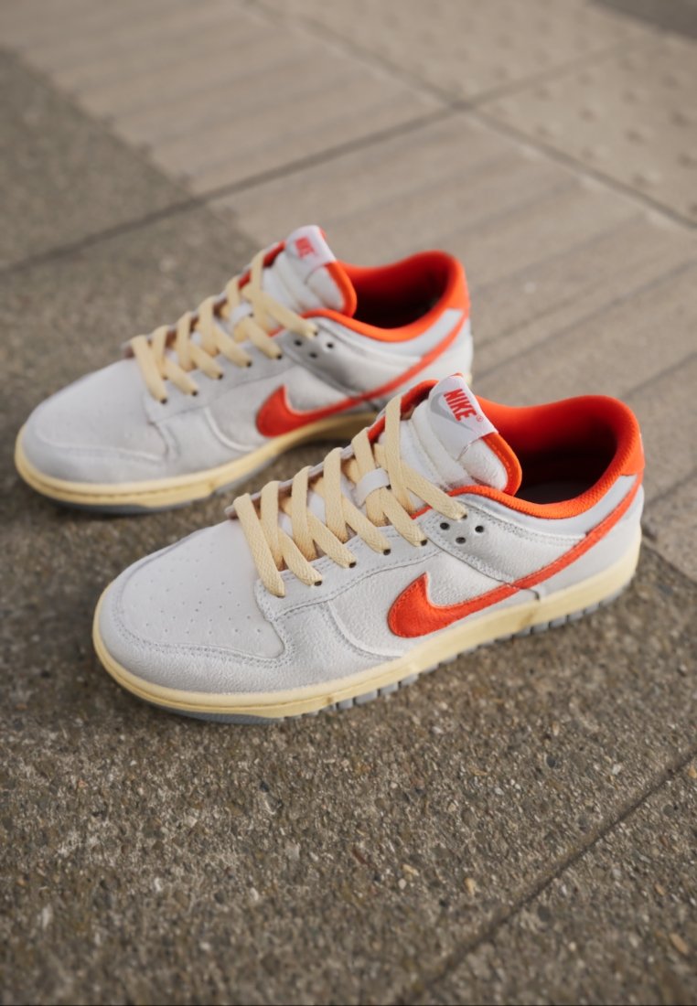 Sneaker Nike bianchi e arancioni con lacci beige poggiati su una superficie pavimentata esterna testurizzata.