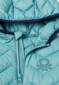 Chaqueta acolchada color teal con capucha, con ribetes en azul marino, cremallera frontal y un logo bordado. La tela tiene una textura suave y brillante.