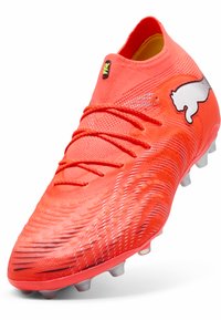 Punainen jalkapallokengän oikea puolisko, jossa on teksturoitu päällinen, valkoinen logo, nauhat ja muotoiltu pohja, jossa on useita nastoja pitoon.