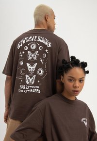 Two people wearing brown t-shirts with white mystical butterfly and celestial designs; one faces forward, the other shows back text and graphics.