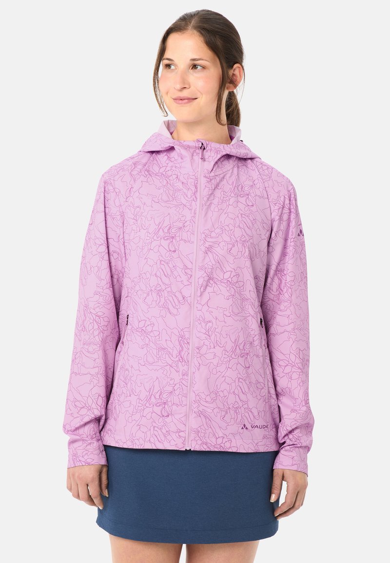 Giovane donna che indossa una giacca con cappuccio color viola chiaro con motivo a zip e una gonna blu scuro, che guarda leggermente di lato con un sorriso dolce.