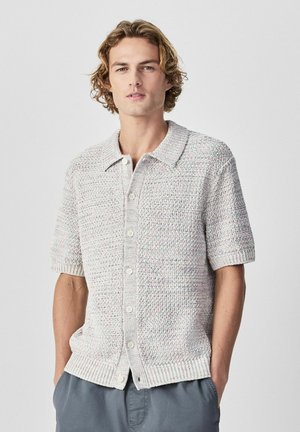 Noor mees laineliste blondide juustega kannab lühikeste varrukatega heledat halli tekstuuriga nööbitavat kardigani ja sinakashalli värvi pükse, käed taskutes.