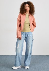 Veste courte rose clair avec passepoil blanc, haut côtelé olive, jean large déchiré, et baskets blanches. Cheveux bouclés, posture debout.