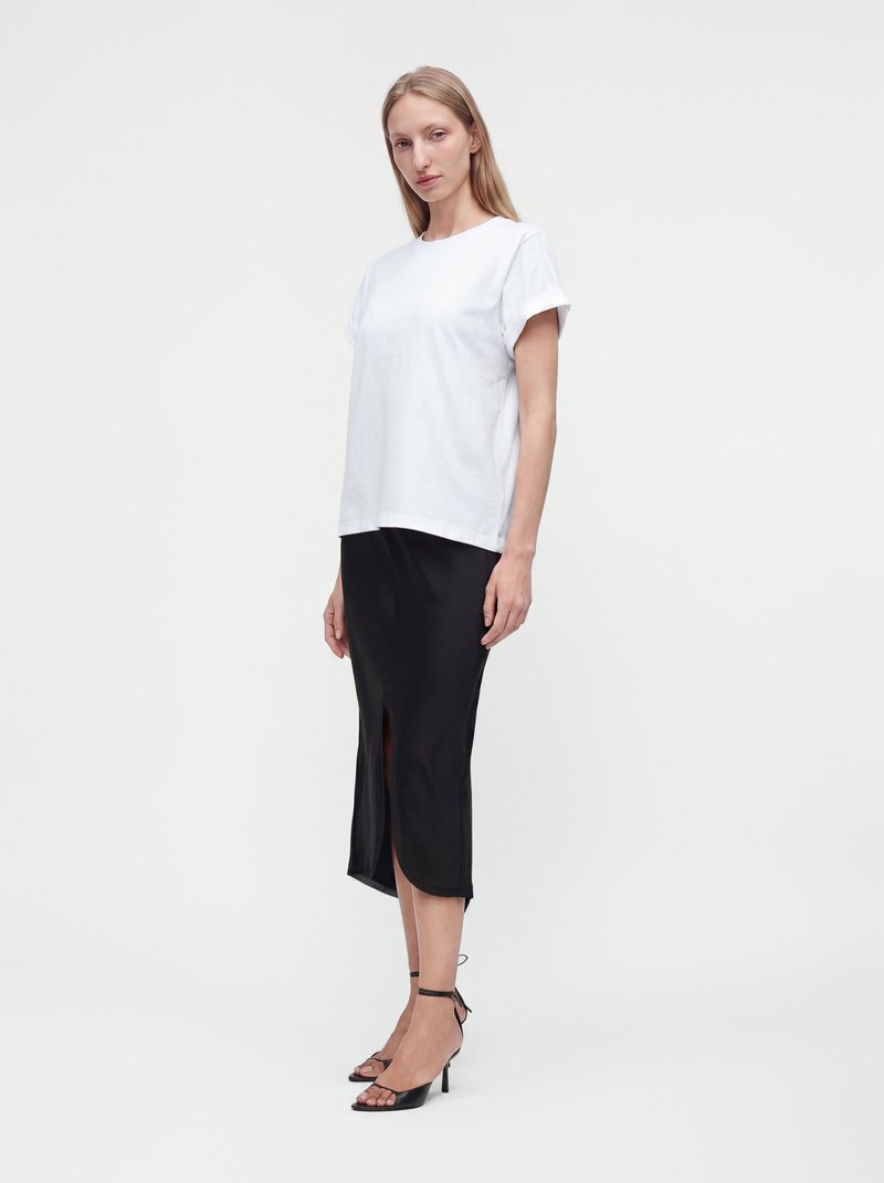 Young woman stands wearing a white short-sleeve top, black mid-length skirt with front slit, and black ankle-strap heeled sandals on a plain background.