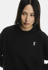 Fekete pulóver puha anyagból, kisebb fehér logóval a mellrészen, kerek kivágással, laza fazonnal és hosszú ujjal.