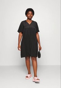 Vestido preto com mangas curtas e fluídas e decote em V, apresentando um corte solto. Usado com sandálias de plataforma rosa e segurando uma pequena bolsa preta.