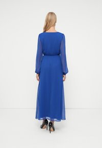 Mulher com cabelo loiro comprido, vestindo um vestido midi azul royal de manga longa e transparente, e sapatos de salto alto pretos, de pé, virada de costas.