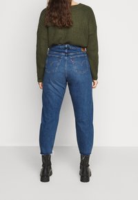 Vysoké modré džíny s uvolněným střihem, zakončené třepením, s zadními kapsami, které jsou v kombinaci s olivově zeleným strukturovaným svetrem.