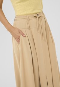 Beige brede benbukser med en tætsiddende talje, prydet med et bælte og læg. Fremstillet af glat stof med sidelommer.