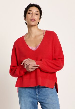 Frau mit kurzem lockigem Haar, die einen locker sitzenden roten V-Ausschnitt-Pullover über einem rosa Spitzen-Top und blauen Jeans trägt, vor einem einfarbigen Hintergrund steht.