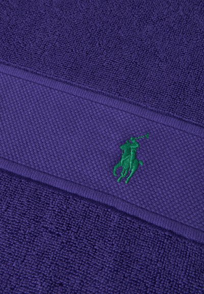 Μωβ πετσέτα με υφή και πράσινο κεντητό λογότυπο ενός παίκτη πόλο πάνω σε άλογο που κρατάει ένα μαλέτι.