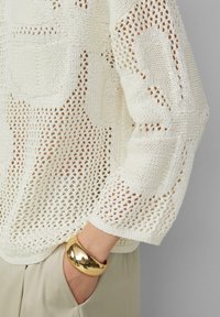 Strickpullover in Creme mit einem lockeren, offenen Webmuster. Verfügt über eine Tasche und lockere Ärmel. Goldene Armreif-Akzent am Handgelenk.