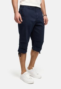 Navy blue cargo shorts made of cotton, featuring knee-length cut, adjustable cuffs with ties, and multiple pockets. Worn with white sneakers.