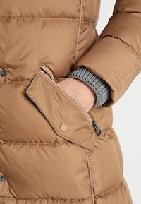 Chaqueta de plumas marrón con textura acolchada, que presenta un bolsillo con cremallera. El puño es de punto gris, añadiendo un contraste al exterior suave.