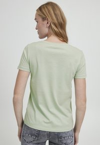 Camiseta verde claro de manga corta con cuello redondo, textura suave y corte relajado, combinada con jeans grises de diseño.