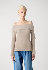 Haut beige côtelé à épaules dénudées avec des manches longues, conçu avec une silhouette ajustée et un tissu texturé. Porté avec un jean bleu à jambes larges.