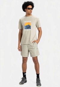 Světle béžové tričko s potiskem hor, kombinované se světlými šortkami a šedými sportovními botami. Doplňky zahrnují černé sluneční brýle a ponožky.