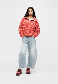 Κόκκινο φλις πουλόβερ με πρότυπο και μισό φερμουάρ, σε συνδυασμό με ανοιχτό μπλε τζιν παντελόνι φαρδιάς γραμμής και κόκκινα αθλητικά παπούτσια με μαύρες λεπτομέρειες.