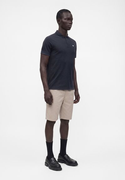 Homme debout portant un polo noir, un short beige, des chaussettes noires mi-mollet et des mocassins noirs sur un fond blanc uni.