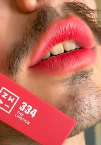 Czerwona szminka o matowym wykończeniu, nałożona na pełne usta; obok widoczny jest pudełko z napisem "ZW 334 THE LIPSTICK" w wyraźnym białym tekście.
