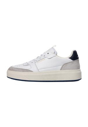 Witte lage sneaker met grijze suède accenten, dikke crèmekleurige zool, marineblauwe hieltab en gouden "CRUYFF" branding aan de zijkant.