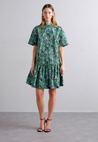 Abito verde a stampa floreale con maniche corte, chiusura con bottoni e orlo a strati con volant. Indossato con tacchi neri con cinturino.
