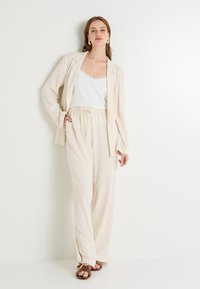 Blazer de lino color beige claro sobre una camiseta blanca, combinado con pantalones de pierna ancha que tienen una cinturilla elástica. Sandalias planas marrones.