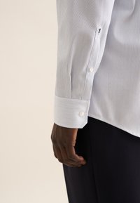Wit en lichtblauw gestreept overhemd met lange mouwen en knoopmanchetten. De manchetten hebben twee knopen en een klein geborduurd detail nabij de naad.