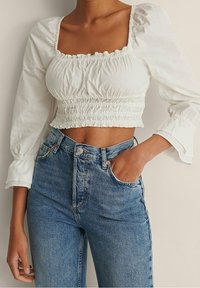 Kobieta w białym, marszczonym topie w stylu crop z bufiasnymi, długimi rękawami oraz w niebieskich, wysokich dżinsach, z ręką w kieszeni.