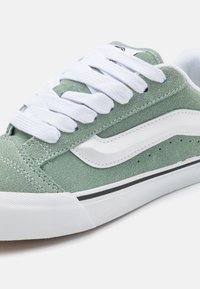 Grønn semsket sneakers med hvite snørebånd, hvit lærstripe, svarte detaljer og teksturert gummisåle; har sømdetaljer og perforeringer.