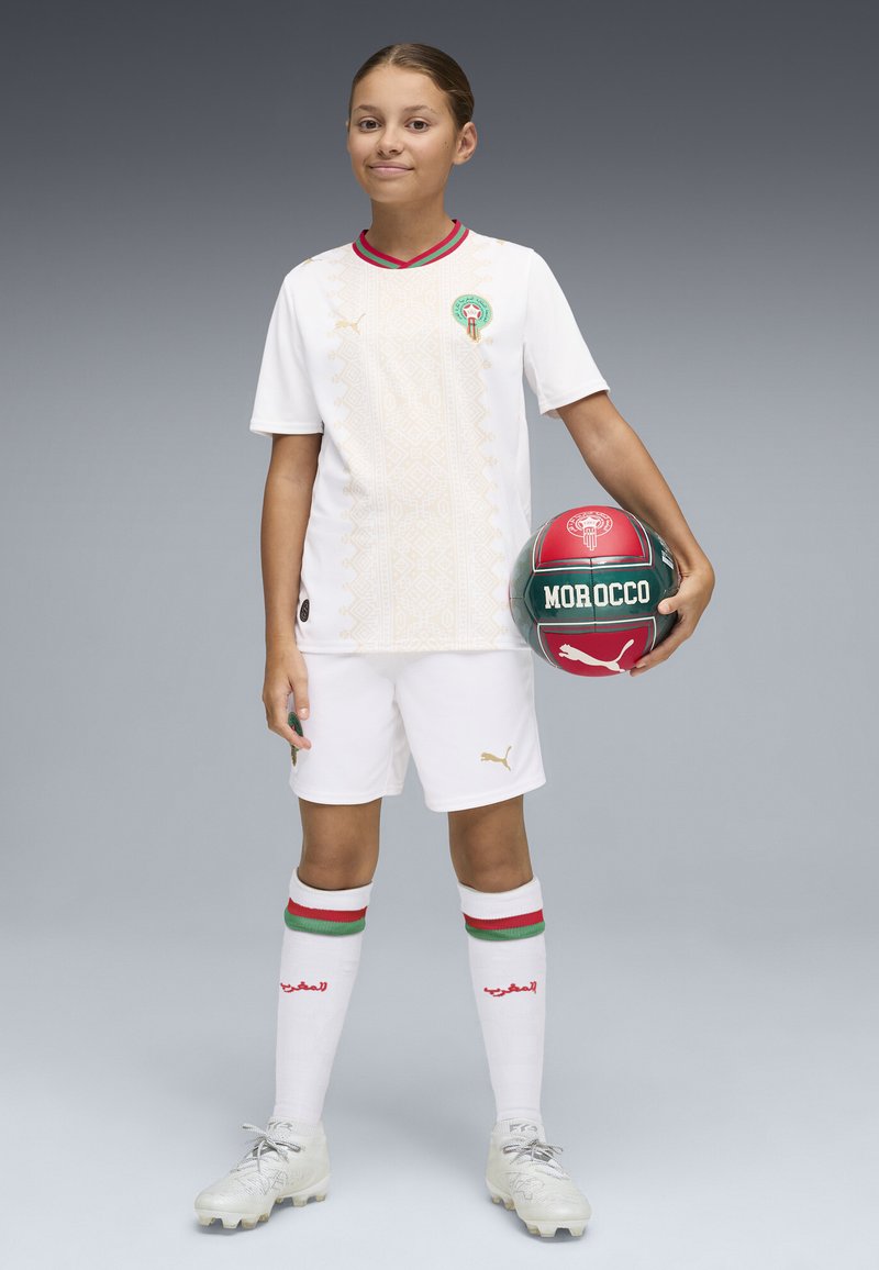 Jeune joueur de football portant le maillot blanc de l'équipe nationale du Maroc, tenant un ballon de football rouge et vert avec le texte "Maroc".