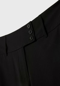 Zone de la taille d'un pantalon noir montrant trois boutons noirs sur la fermeture latérale et des passants de ceinture sur un fond uni clair.