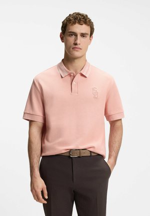 Uomo con polo rosa chiaro a maniche corte con logo ricamato, pantaloni scuri e cintura marrone, in piedi con una mano in tasca.