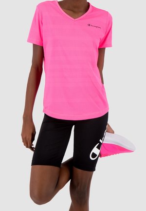 Persona atletica che si allunga una gamba, indossando una maglietta rosa acceso a maniche corte e pantaloncini neri con un logo bianco sul fianco.