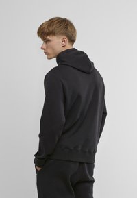 Hanorac negru cu tiv curbat, mansete ribbate și glugă cu șnur. Textură de țesătură netedă, design minimalist și croială lejeră.
