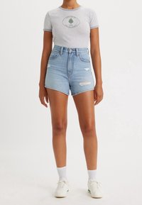 T-shirt grigio chiaro con logo verde, abbinato a pantaloni corti in denim a vita alta di colore azzurro chiaro, con dettagli strappati e orlo sfrangiato.