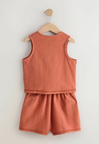 Rustfarvet ærmeløst sæt med top og shorts med elastisk talje hængende på en træbøjle mod en ensfarvet baggrund.