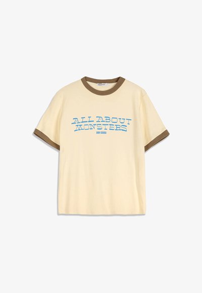 T-shirt beige à manches courtes avec bordure marron au col et aux manches, avec un texte bleu "ALL ABOUT MONSTERS" sur le devant.