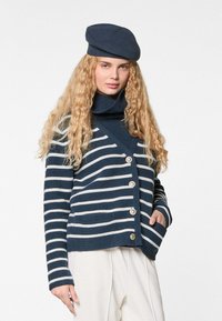 Marineblauwe en witte gestreepte cardi met een kraag, met knopen aan de voorkant en zakken. Gebreide textuur, gecombineerd met een bijpassende beret.