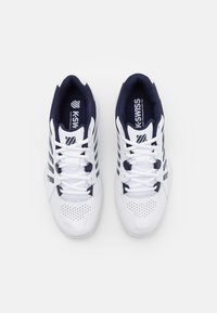 White athletic shoes with navy accents, featuring mesh and synthetic materials, lace-up design, perforated toe box, and brand logo on tongue.