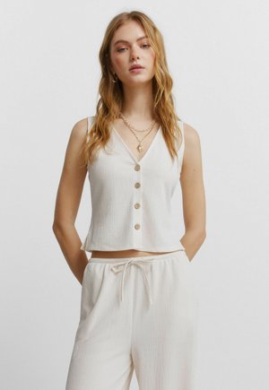 Vrouw met lang blond haar, gekleed in een mouwloos wit hemd met knopen en bijpassende broek met trekkoord, staande voor een effen achtergrond.