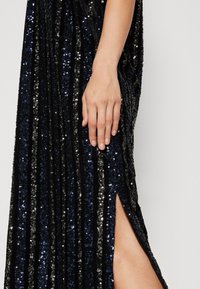 Svart paljettklänning med vertikala veck och blå och silverfärgade detaljer. En hög slits vid låret avslöjar hud bredvid en hand som vilar på tyget.