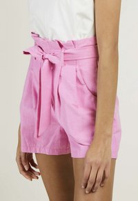 Shorts en lin rose présentant une taille haute, un devant plissé et une ceinture à nouer. Le design inclut des poches latérales et une texture lisse.